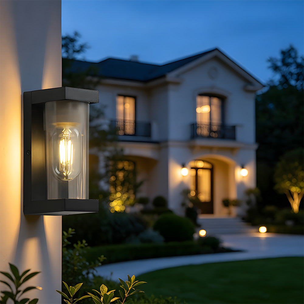 A modern outdoor wall light fixture mounted on a house exterior at dusk, casting a warm glow on the wall. The fixture has a matte black rectangular frame and a clear glass shade housing an exposed filament bulb.